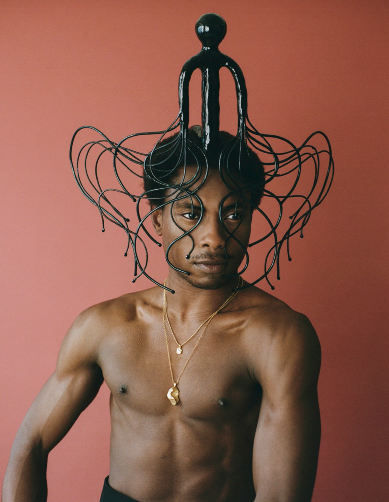Model wearing black sculptural headpiece with two gold necklaces